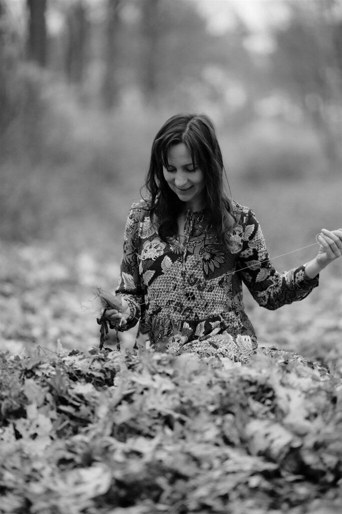 Monika Bakos sewing on the forest ground, holding autumn leaves, black‑and‑white portrait.